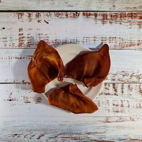 Three pigs ears dog chews on a cream plate on a whitewashed wooden board