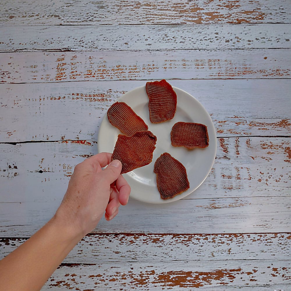 A hand holding a piece of smoked ostrich dog treat above a cream plate to show its size